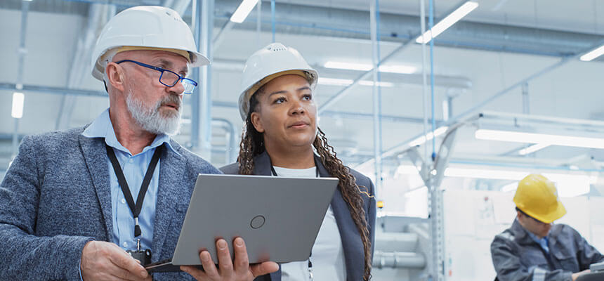 Zwei Mitarbeitende mit Schutzhelmen und Laptop in einer Produktionshalle als Bild für Compliance, Arbeitssicherheit und rechtssichere Prozesse