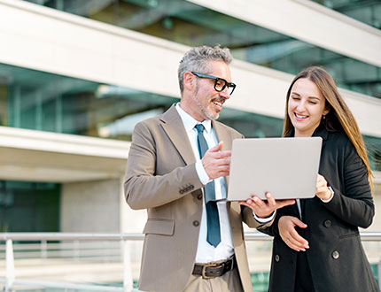 CSRD DNK GRI ZNU Bericht ESRS-konform, älterer Mann und junge Frau am Laptop vor Bürogebäude