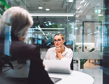 EU Taxonomie Berichterstattung, modernes Büro, gläserne Wände, Frau mit Laptop sitzt Mann gegenüber an Konferenztisch