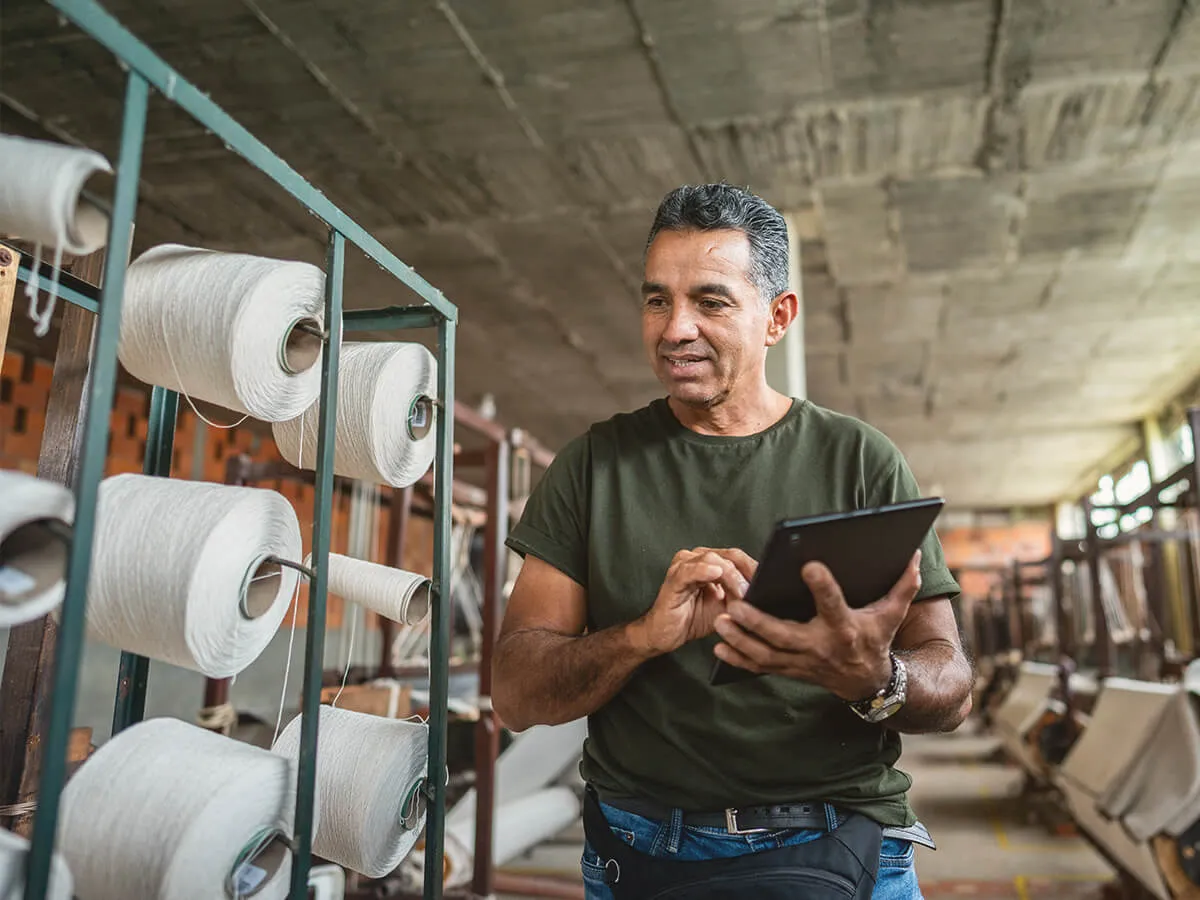 PCF Magazinartikel Bild, globaler Süden, Mann mit Tablet in der Hand geht in einer kleinen Fabrikhalle an großen Stoffspulen vorbei