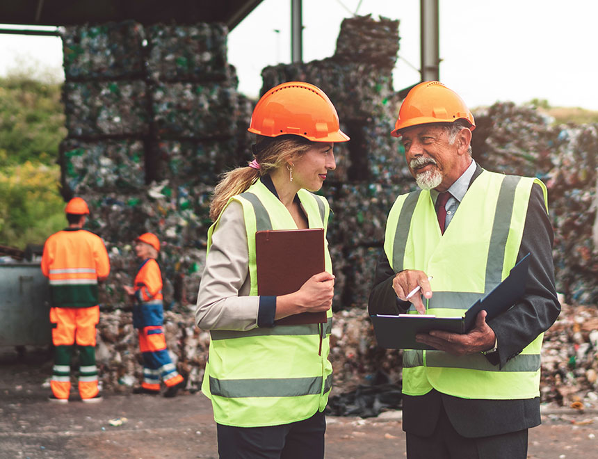 Zwei Mitarbeitende prüfen Unterlagen vor gepressten Kunststoffballen in einer Recyclinganlage.